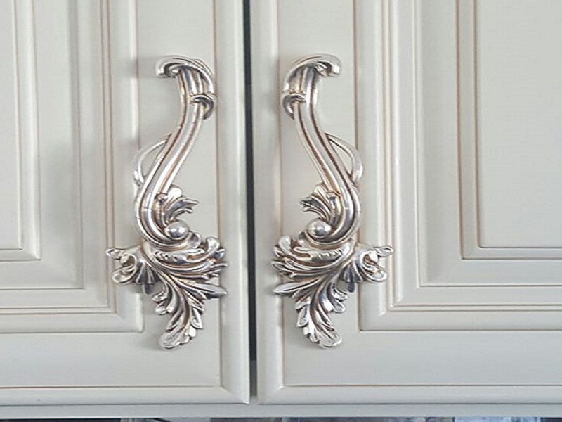 دستگیره کابینت کلاسیک نقره‌ای با طرح منبت‌کاری روی درب سفید قاب‌دار.