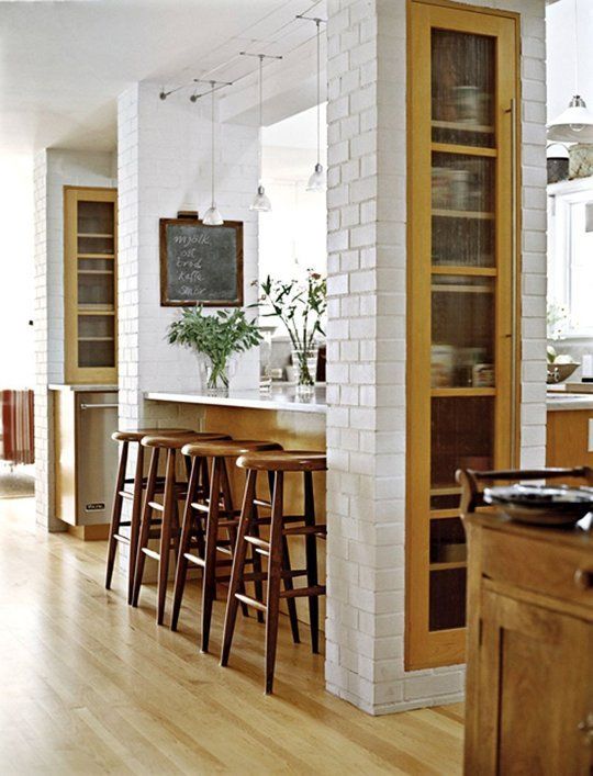 یک آشپزخانه با ستون‌های آجری سفید، کابینت‌های چوبی و صندلی‌های بار چوبی