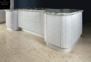 نمای نزدیک از جزیره آشپزخانه با طراحی منحنی، صفحه رویی سنگی و درب‌های شیار‌دار سفید