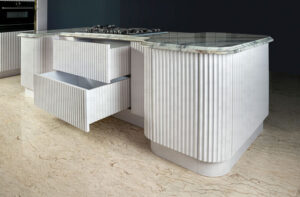 نمای نزدیک از جزیره آشپزخانه با طراحی منحنی، صفحه رویی سنگی و درب‌های شیار‌دار سفید