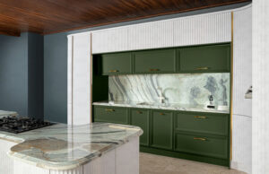 جزیره‌ی آشپزخانه با فرم منحنی و سطح کار سنگی خاکستری، بخشی از کابینت‌های مدرن سفید و سبز