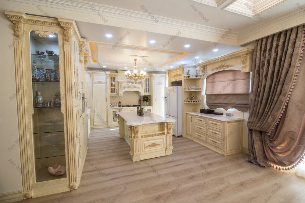 کابینت چوبی کلاسیک آشپزخانه با طراحی منبت‌کاری شده، رنگ استخوانی با پتینه طلایی، جزیره مرکزی و ویترین شیشه‌ای