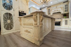 کابینت چوبی کلاسیک آشپزخانه با طراحی منبت‌کاری شده، رنگ استخوانی با پتینه طلایی، جزیره مرکزی و ویترین شیشه‌ای