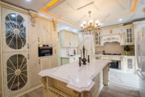 کابینت چوبی کلاسیک آشپزخانه با طراحی منبت‌کاری شده، رنگ استخوانی با پتینه طلایی، جزیره مرکزی و ویترین شیشه‌ای