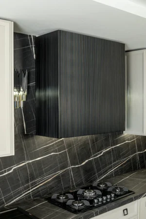 هود آشپزخانه در طراحی نئوکلاسیک، با ظاهری مدرن و خطوط مینیمالیستی، که بر روی پس‌زمینه‌ای از کاشی‌های مشکی و طرح‌های سفید قرار دارد. این هود با طراحی ساده و شیک خود به‌خوبی با فضای آشپزخانه‌های نئوکلاسیک هماهنگ می‌شود و جلوه‌ای مدرن و در عین حال کلاسیک به محیط می‌بخشد